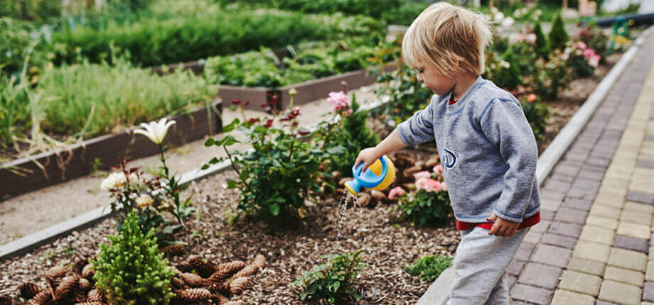 A boy with long blond hair is watering flower beds in the summer 