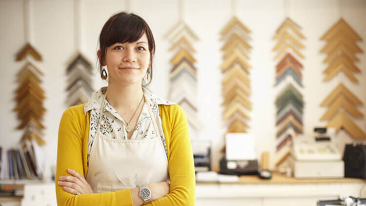 Female business owner stood in her framing studio wearing a yellow cardigan with arms folded looking into the camera
